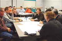 A group of people sit around tables in a meeting room, discussing and writing on large sheets of paper.