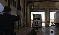 An officer signals a white semi-truck to enter an inspection bay.