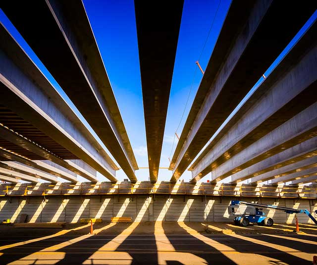 View from beneath a bridge under construction, showing parallel concrete beams and a blue sky above, with a construction lift vehicle on the right.