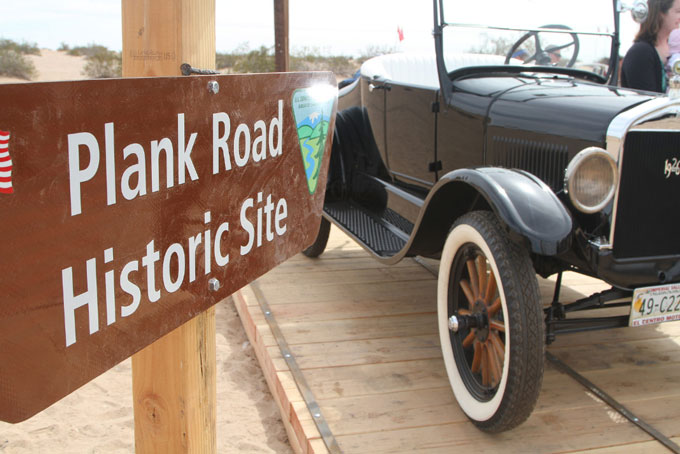 A vintage car parked on wooden planks near a Plank Road Historic Site sign in a desert area.
