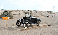 A vintage black car is displayed on a wooden platform in a sandy desert area with a sign nearby.