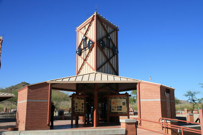 A brick and metal visitor center with a tall tower structure, information boards, and a clear blue sky in the background.