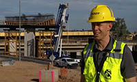 A construction worker in a yellow safety vest and hard hat stands at a building site with cranes, vehicles, and an overpass structure in the background.