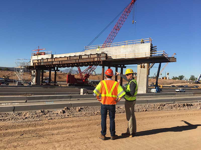 Two construction workers in safety gear stand on a dirt road, observing a partially built bridge with cranes and equipment on a clear day.