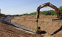 Workers and an excavator build a canal or ditch, lining it with material on a sunny day.