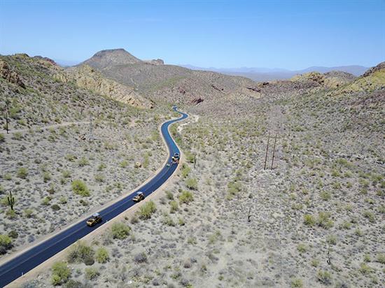 A winding road with several vehicles passes through a dry, rocky desert landscape under a clear blue sky.
