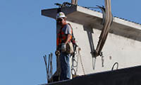 Construction worker in safety gear stands on a beam near large concrete structure under blue sky.