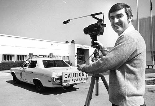Black and white image of a man with a camera standing in front of a car that has a "Caution: Skid Research" sign on the bumper