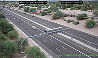 A highway with several vehicles traveling in both directions, surrounded by desert landscaping and trees, viewed from an overhead traffic camera.