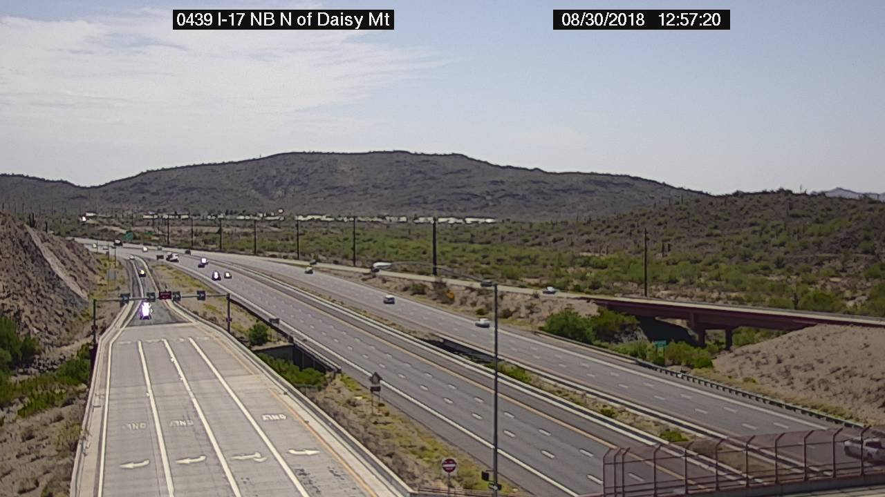 A mostly empty highway near mountains, with a few cars and clear skies, seen from a traffic camera.