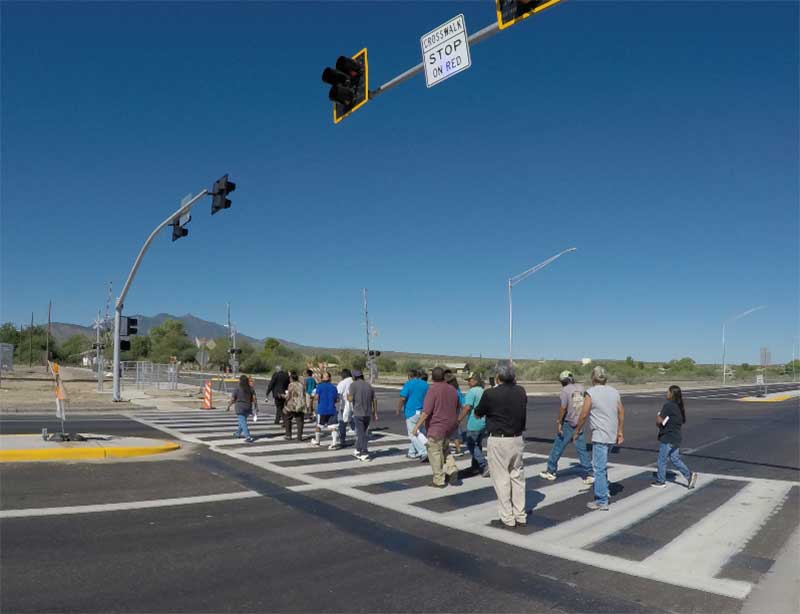 A group of people crosses a wide street at a crosswalk under a clear blue sky and traffic lights.