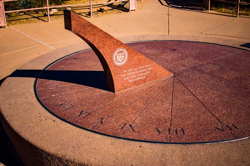 Large stone sundial with Roman numerals, an upright triangular gnomon, and an inscribed text in the center, set in a paved outdoor area.