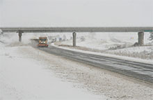 A snowplow clears a snow-covered road beneath an overpass on a wintry day.