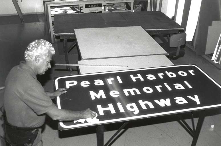 Black and white photo of a man working on a Pearl Harbor Memorial Highway sign.