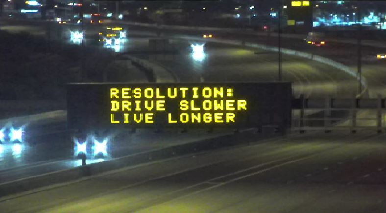 An electronic highway sign displays the message: RESOLUTION: DRIVE SLOWER LIVE LONGER to encourage safe driving at night.
