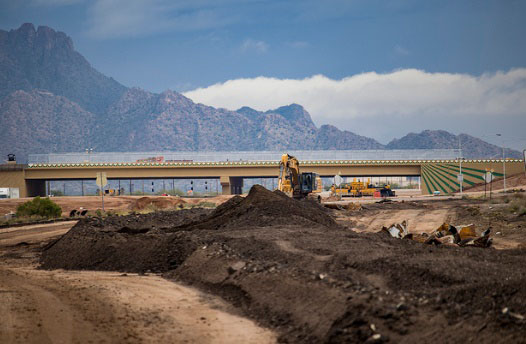 Excavators work on a dirt construction site near a bridge with mountains in the background.