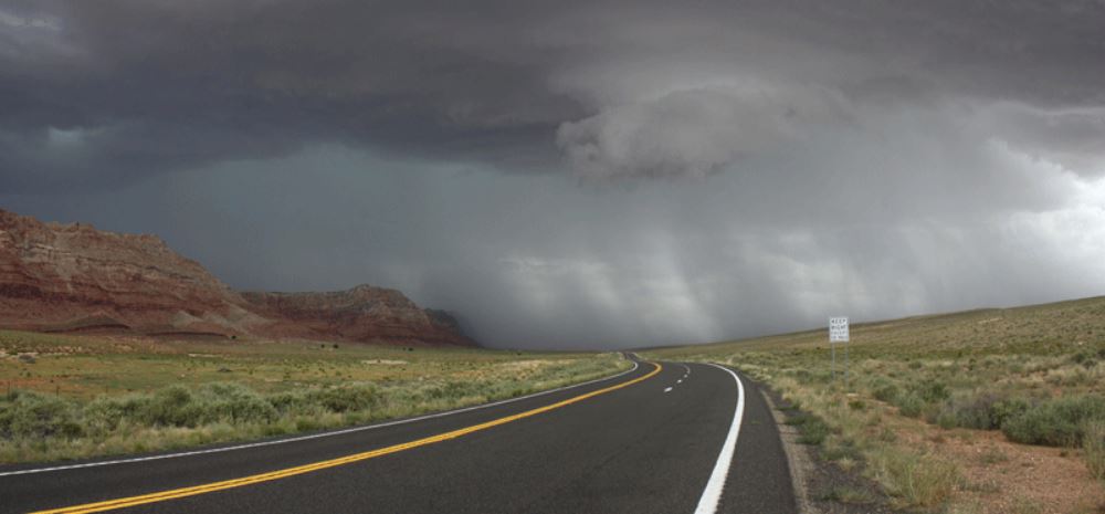 A winding road leads to distant mountains under a dark, stormy sky with rain falling in the distance.