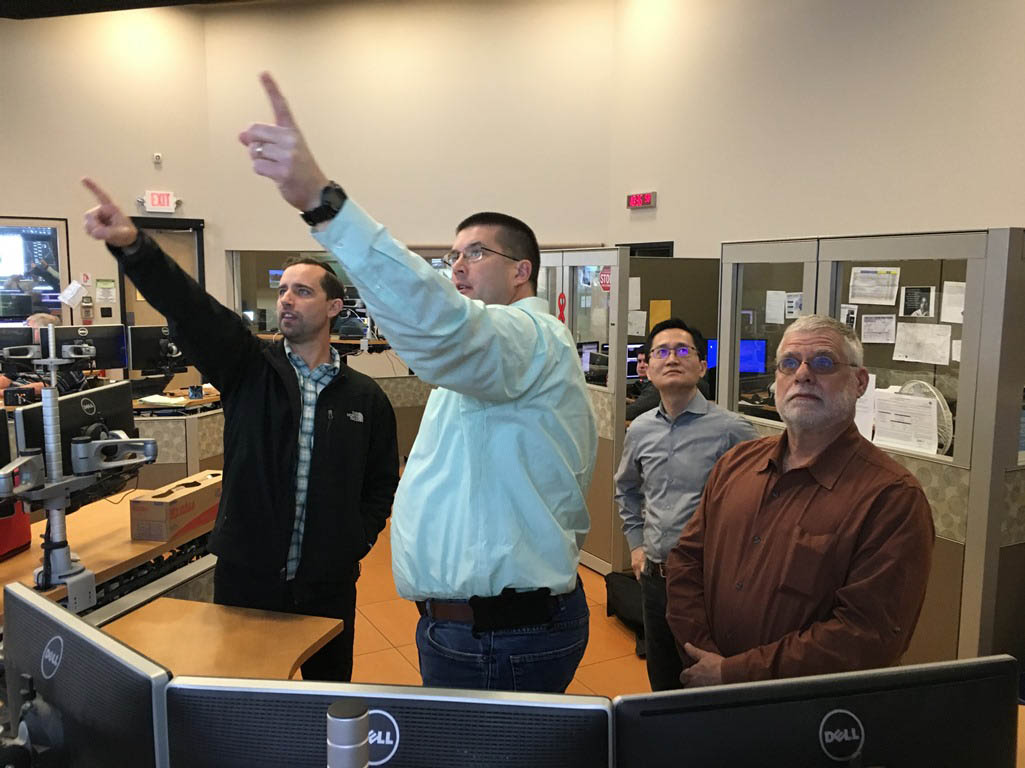 Four men stand in an office, two of them pointing at monitors, while the others observe. Computer screens and office equipment are visible.