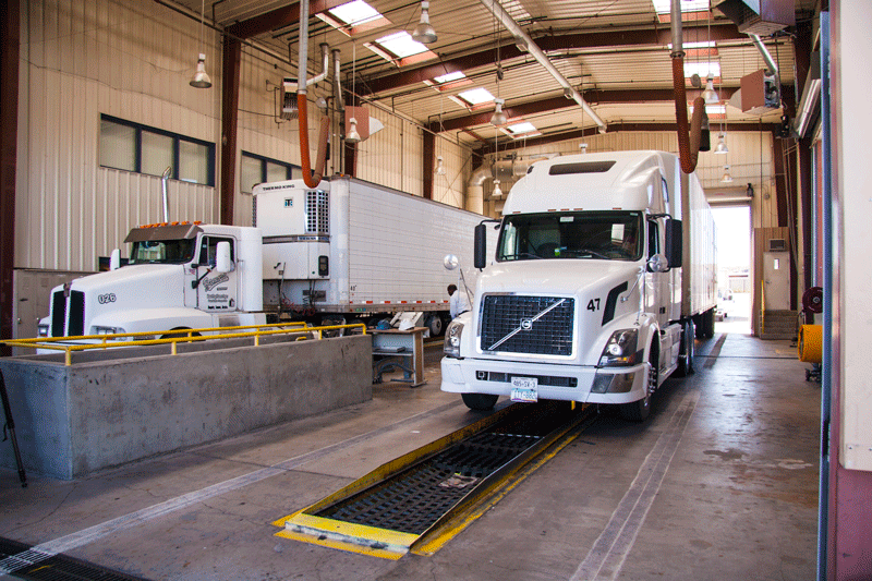 Two white semi-trucks inside a large ADOT inspection facility with high ceilings and overhead lights.