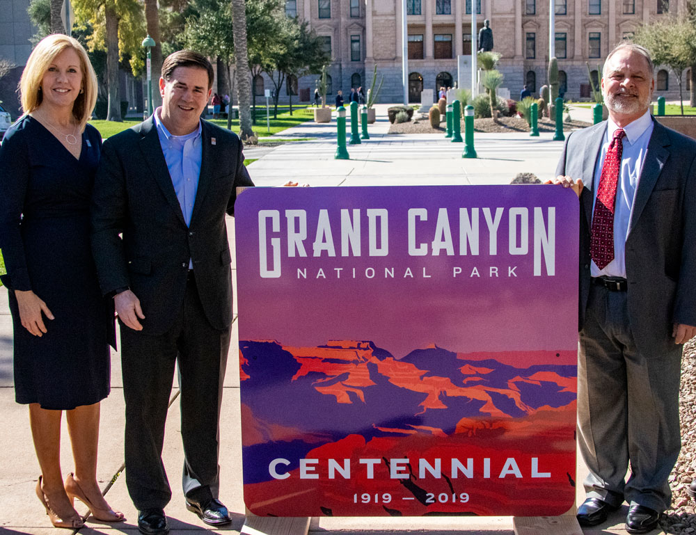 Three people stand outdoors holding a Grand Canyon National Park Centennial sign, 1919-2019.