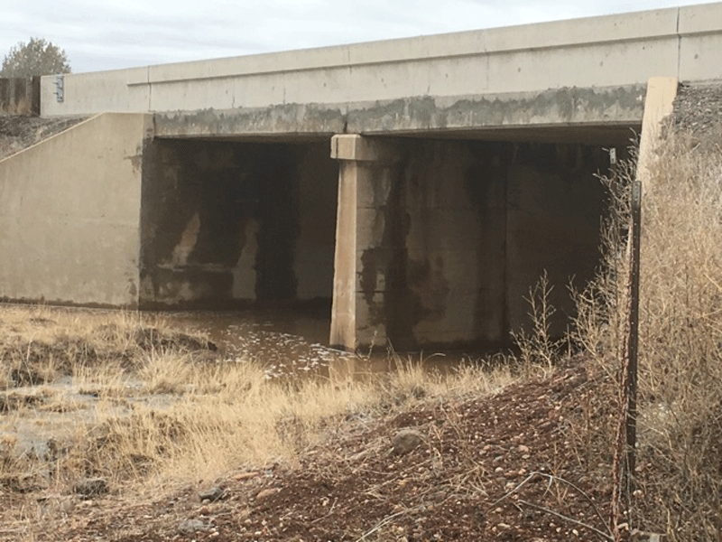 Concrete bridge over a shallow stream with dry grass and rocks in the foreground; stained walls indicate water flow levels.
