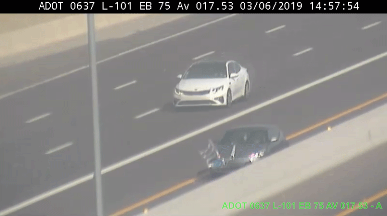 Traffic camera view of two cars traveling on a multi-lane highway; one white car in the lane, one darker car with flags on the shoulder. A ladder appears to be caught on the dark car's hood.