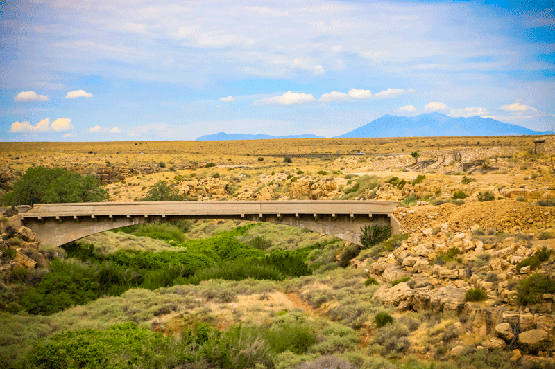 A concrete bridge spans a green, brush-filled arroyo in a dry desert landscape with distant mountains and a partly cloudy sky in the background.