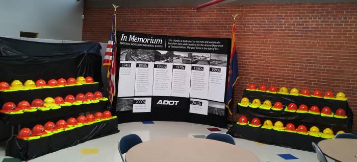 A display of orange and yellow hard hats honors fallen highway workers, with a memorial banner and US flags in the background.