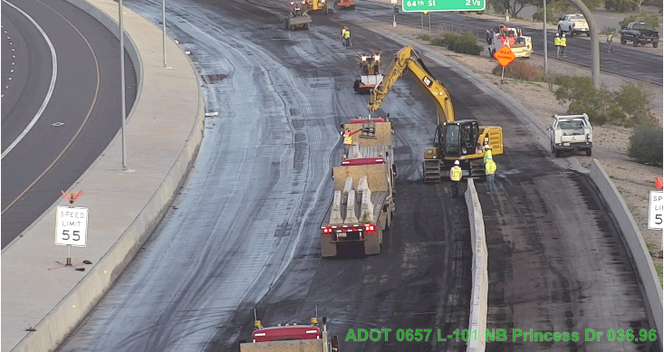 Construction workers and machinery lay concrete barriers on a multi-lane highway with traffic signs and equipment visible along the road.