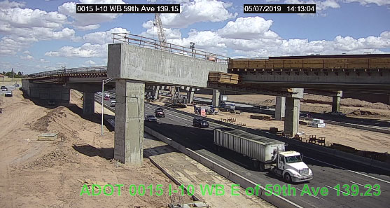 A white semi-truck drives under a concrete overpass under construction, with cranes and unfinished sections visible; blue sky with scattered clouds above.