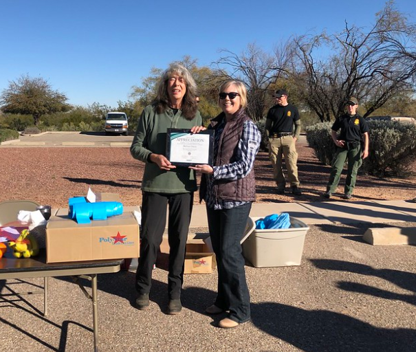 Two women stand outdoors, one holding a certificate of appreciation. Supplies and boxes are on a table nearby, with several people standing in the background.