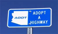 Blue road sign reads ADOT Adopt A Highway with an outline of the state of Arizona and the letters ADOT inside the outline, against a clear blue sky.
