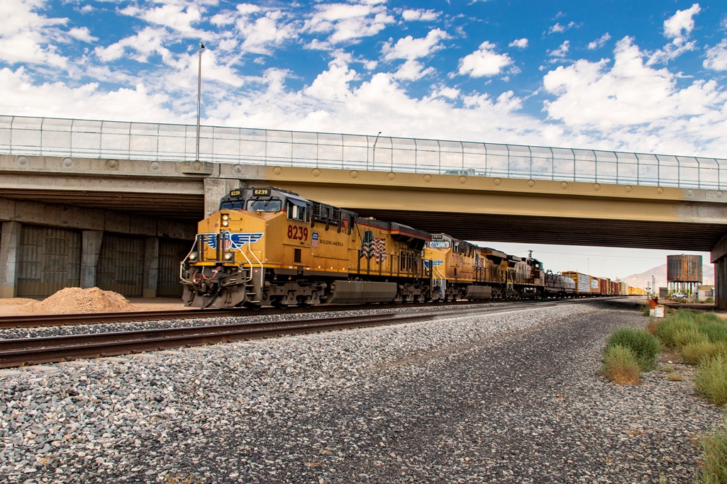 A yellow freight train travels on railroad tracks beneath a concrete overpass, with a blue sky and clouds in the background.