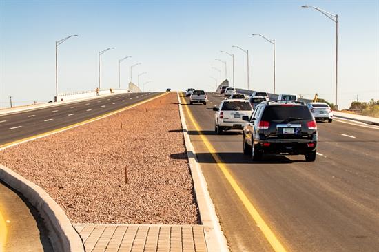 Several vehicles drive up a divided highway overpass on a clear day, with streetlights lining both sides and a rocky median in the center.