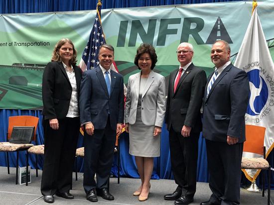 Five people in business attire pose for a group photo in front of a transportation-themed banner and U.S. flags at an official event.
