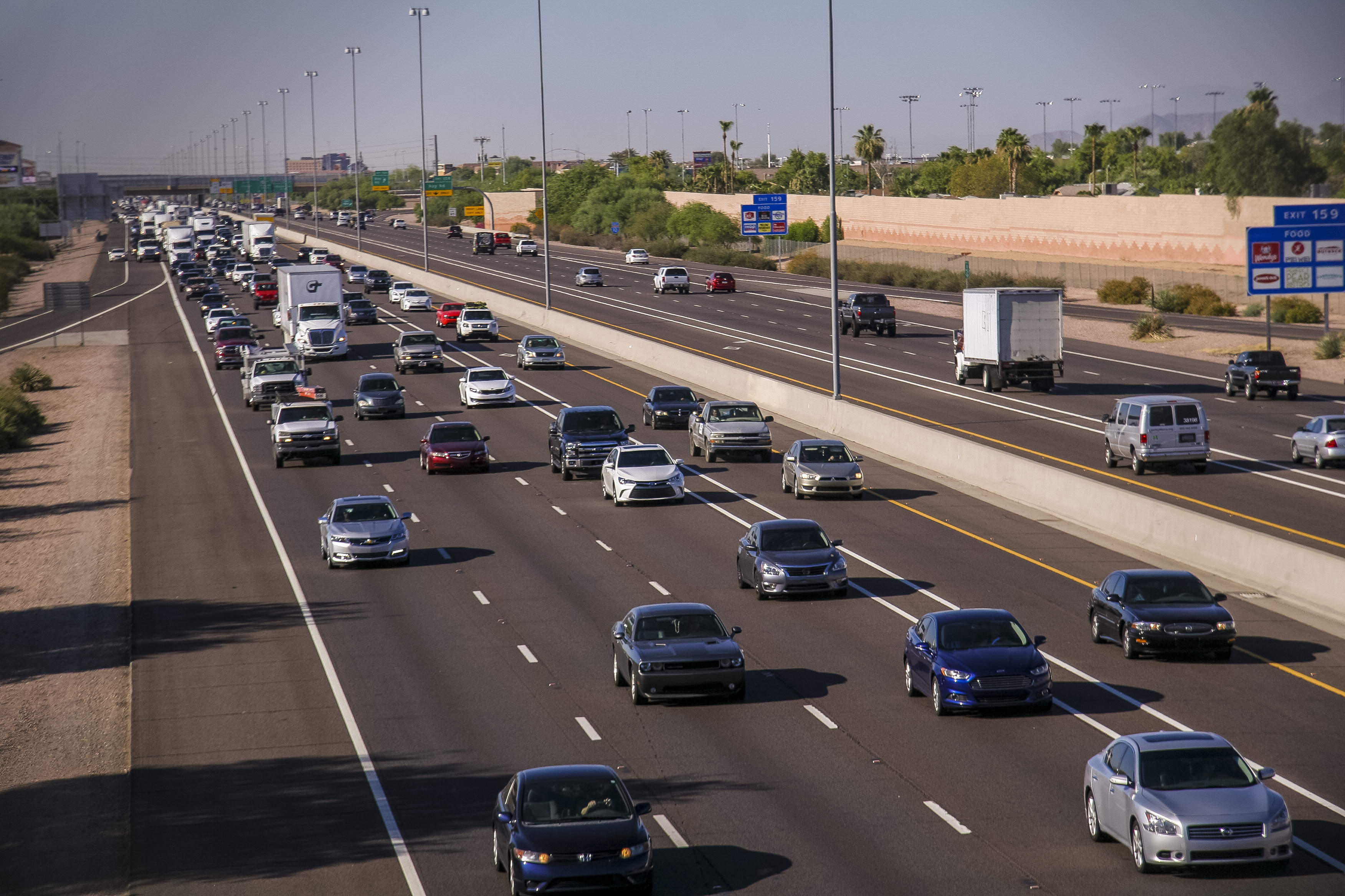 Multiple lanes of vehicles traveling in both directions on a busy highway bordered by concrete barriers and roadside signs under clear skies.