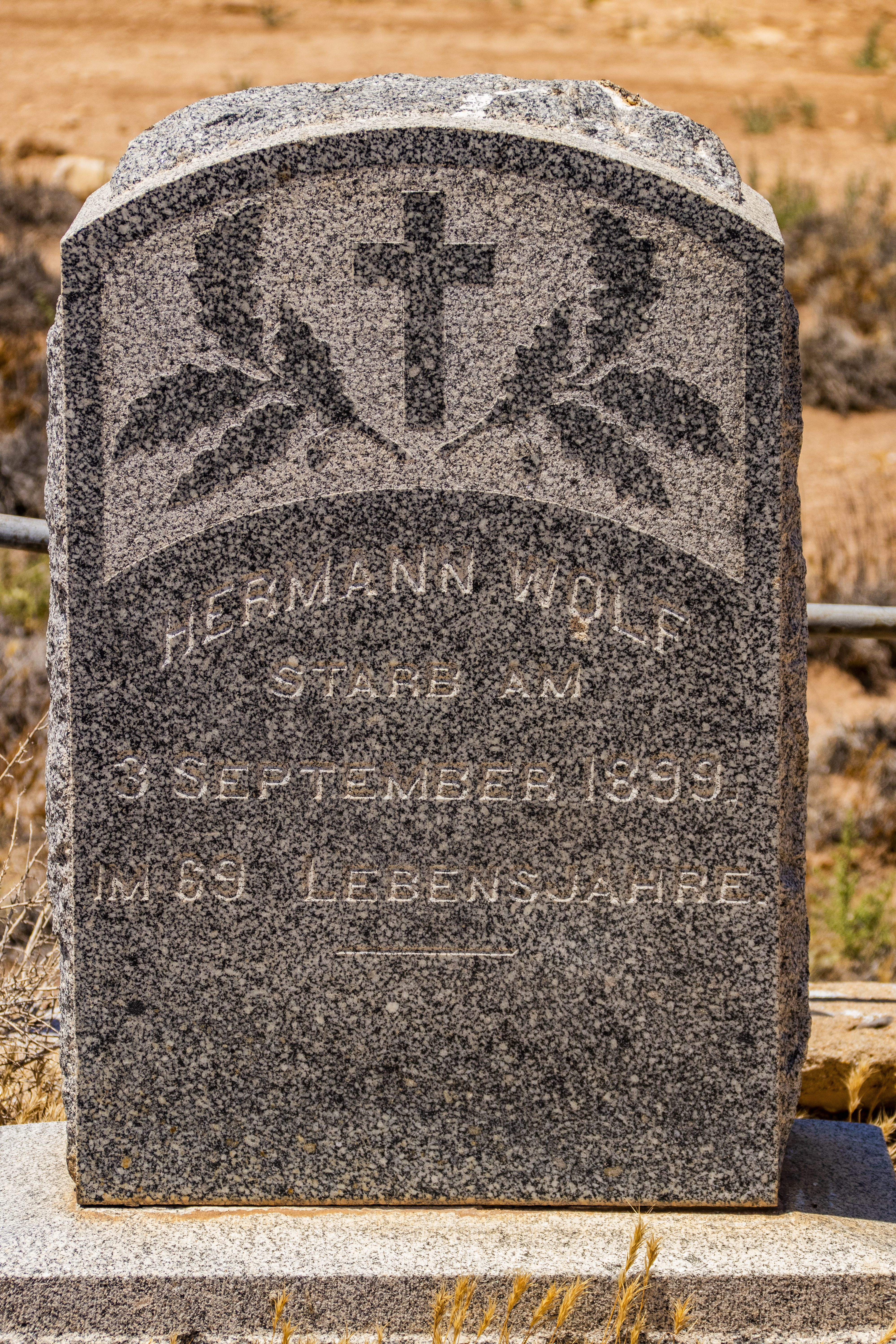 A weathered gravestone curved at the top with a cross and leaves carved into the stone. The name Hermann Wolf is carved into the stone along with birth and death dates.