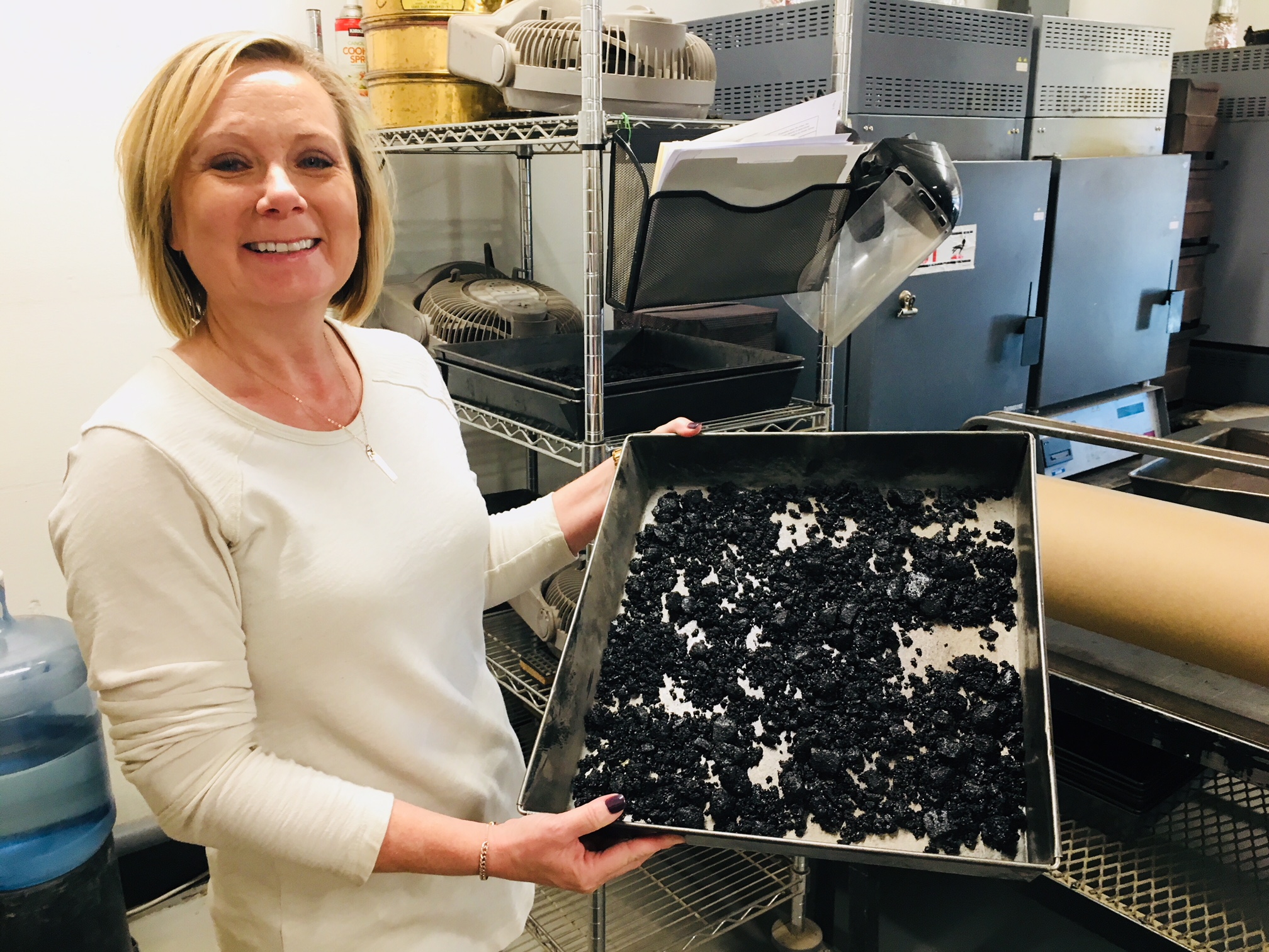 A woman stands in a laboratory holding a metal tray filled with dried black material, with shelves and lab equipment in the background.
