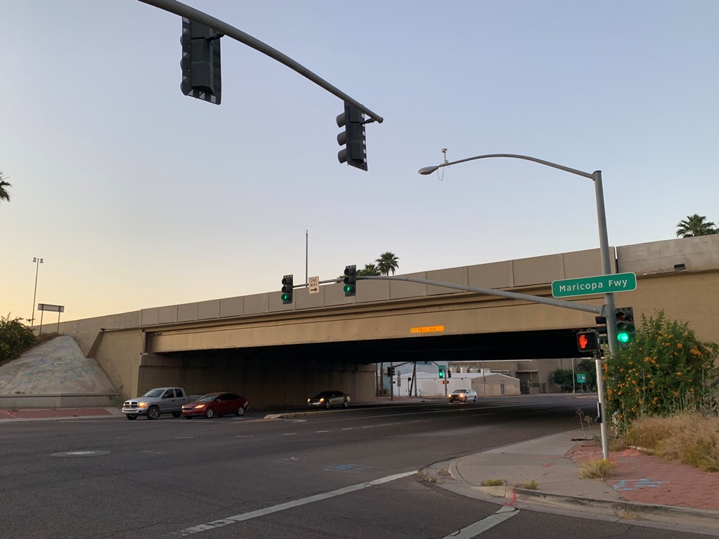 Cars at an intersection under a freeway overpass with a sign reading Maricopa Fwy at dusk.