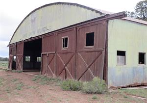 A large, weathered barn with wooden doors and windows, featuring a partially open entrance and faded green metal siding. Shrubs and dirt surround the structure.