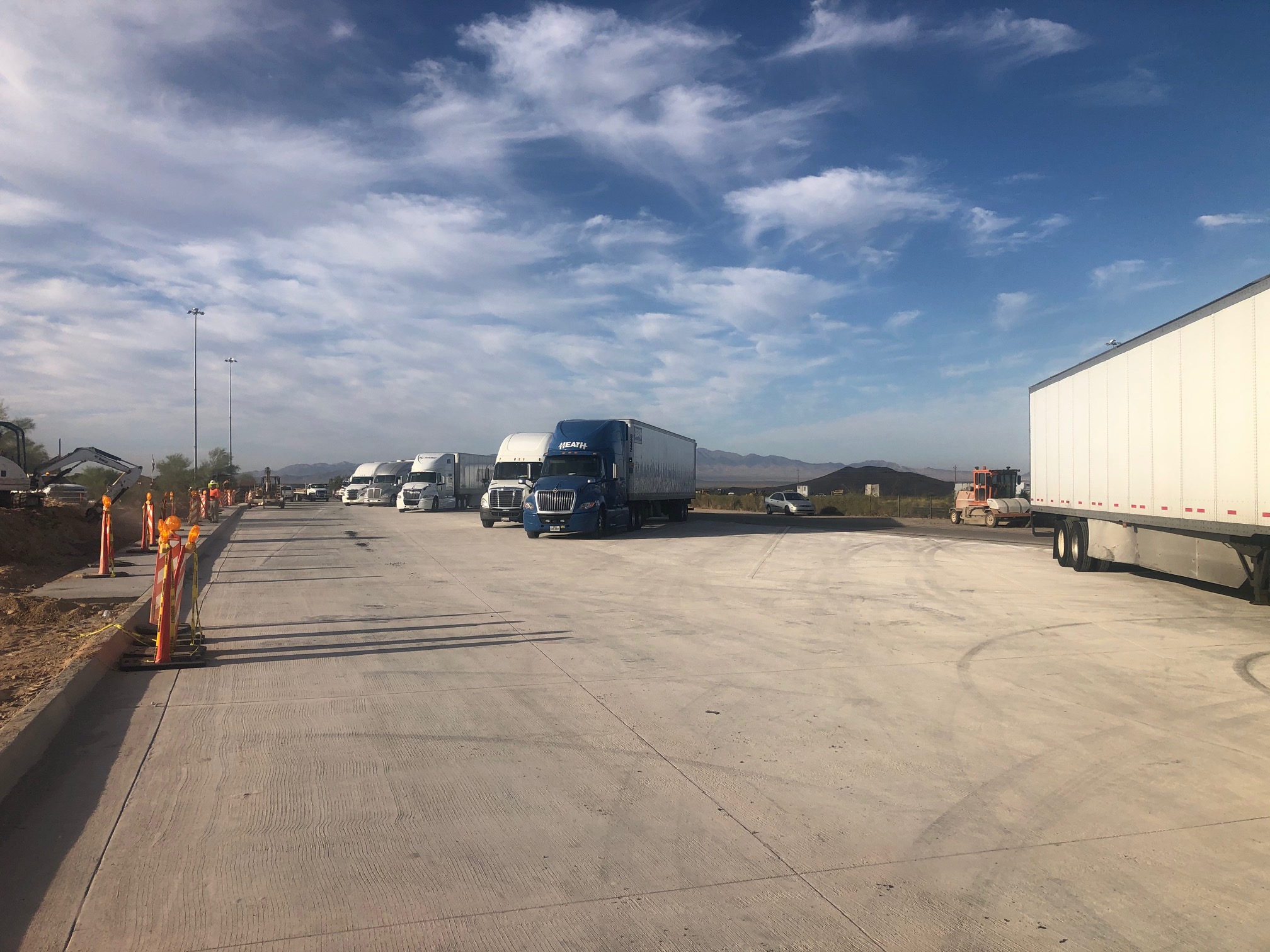 Several semi-trucks parked on a wide concrete lot under a partly cloudy sky with mountains in the distance.