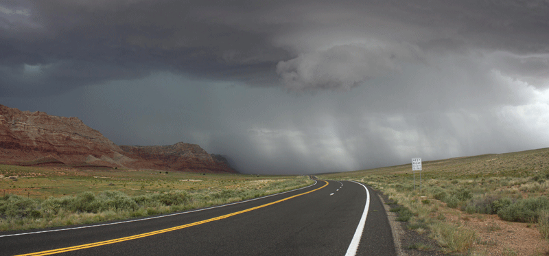 A two-lane highway curves through a desert landscape under dark storm clouds with rain falling in the distance; red rock cliffs appear on the left.