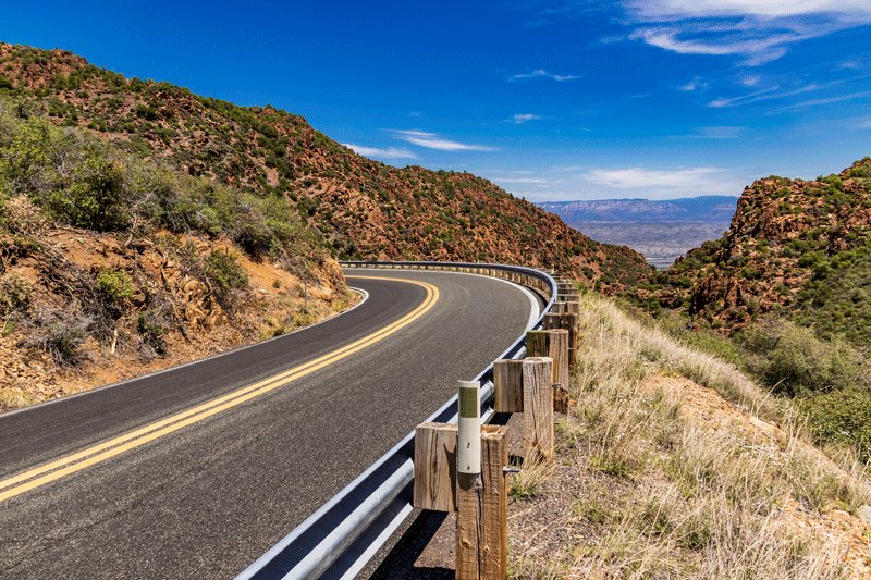 A curving Arizona highway with guardrails surrounded by rocky hills and clear blue sky.