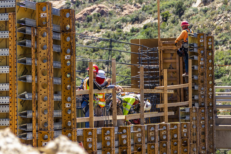 Construction workers in safety gear working on a bridge framework with metal supports and green hills behind.