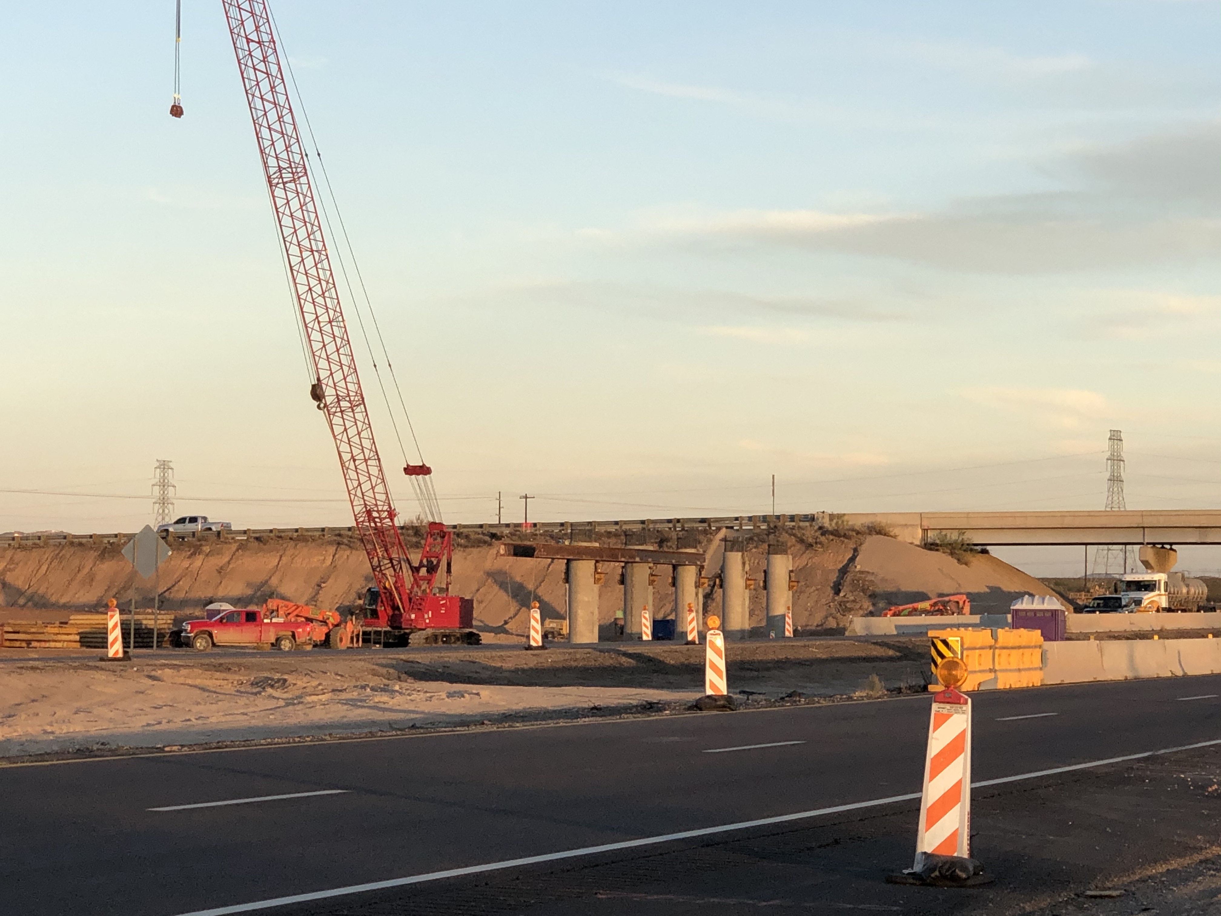 A construction site with a red crane, concrete pillars, sand piles, and caution barriers near a highway under an evening sky.