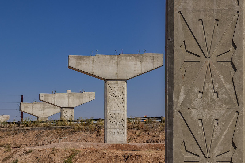 Three unfinished concrete bridge supports stand on a dirt embankment under a clear blue sky, with stylized monarch butterfly patterns visible on the closest pillar.