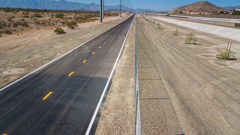 A paved road runs parallel to a canal in a dry, desert landscape with mountains in the distance.