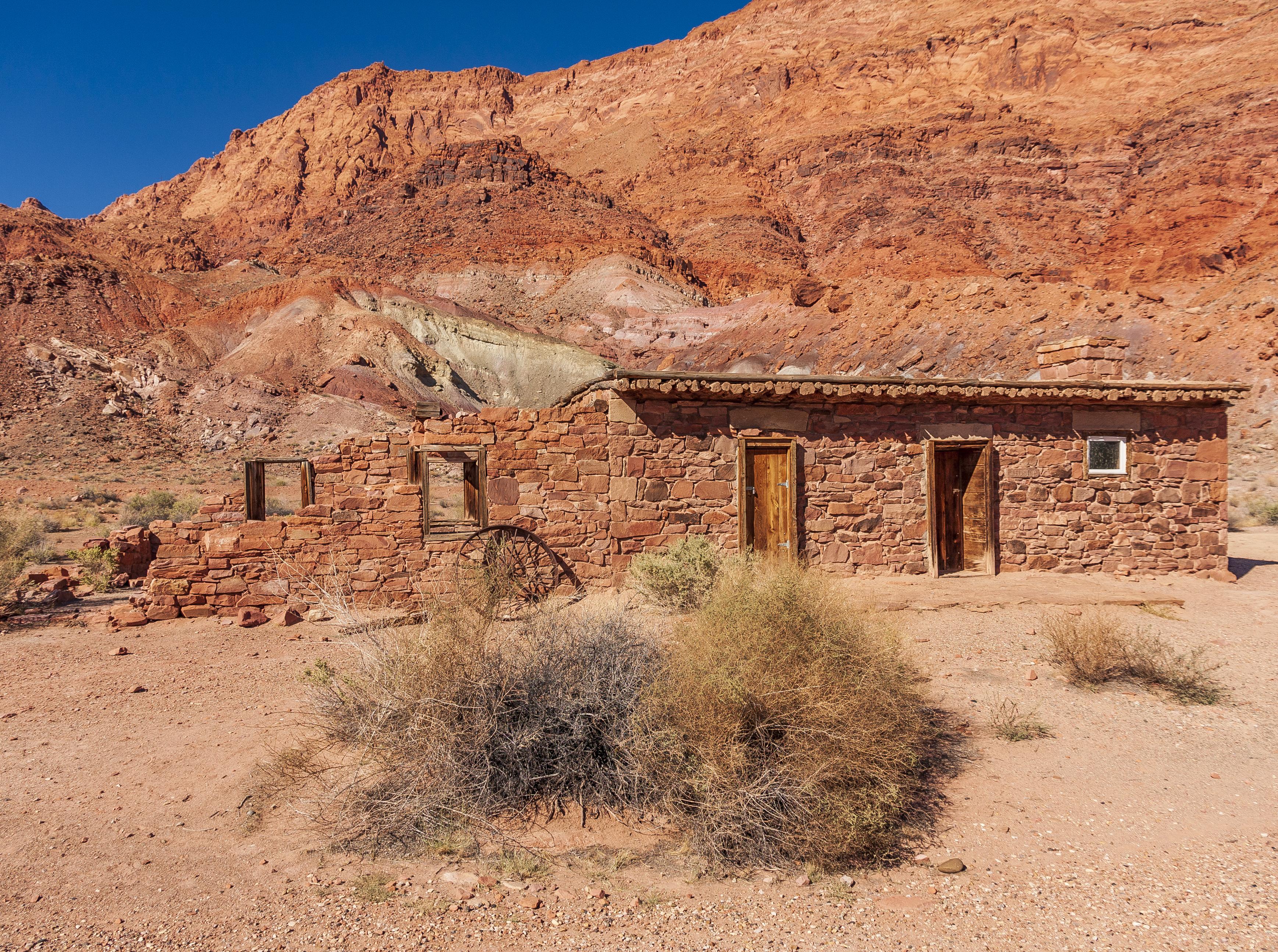 A small, abandoned stone building with wooden doors stands in a dry desert landscape against a backdrop of red rocky hills under a clear blue sky.