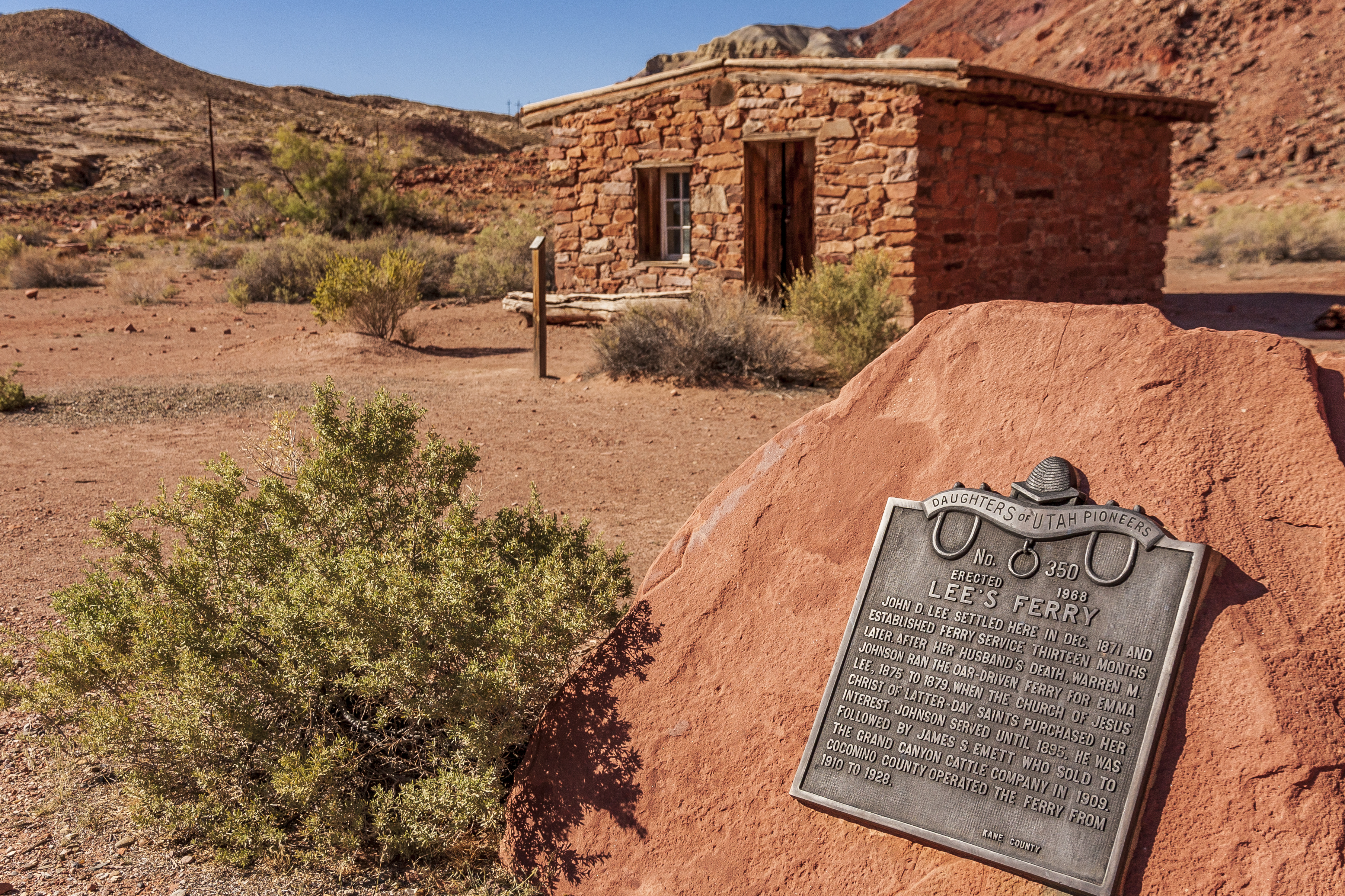 A historic stone building stands in the desert behind a commemorative plaque mounted on a large red rock, surrounded by dry vegetation and hills.