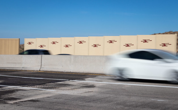 A white car speeds past a beige highway sound wall decorated with red geometric designs.
